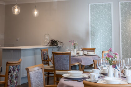 Bright and cheerful eating area in York Manor Care Home, featuring dining tables set with colorful tableware and a small vase with fresh flowers on each table.