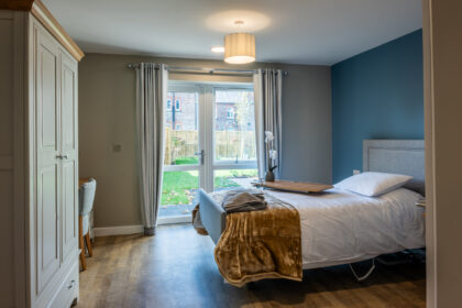 Cozy bedroom inside York Manor Care Home, featuring an inviting bed with soft linens, a bedside table with a lamp, and large windows with open curtains allowing natural light to fill the room.