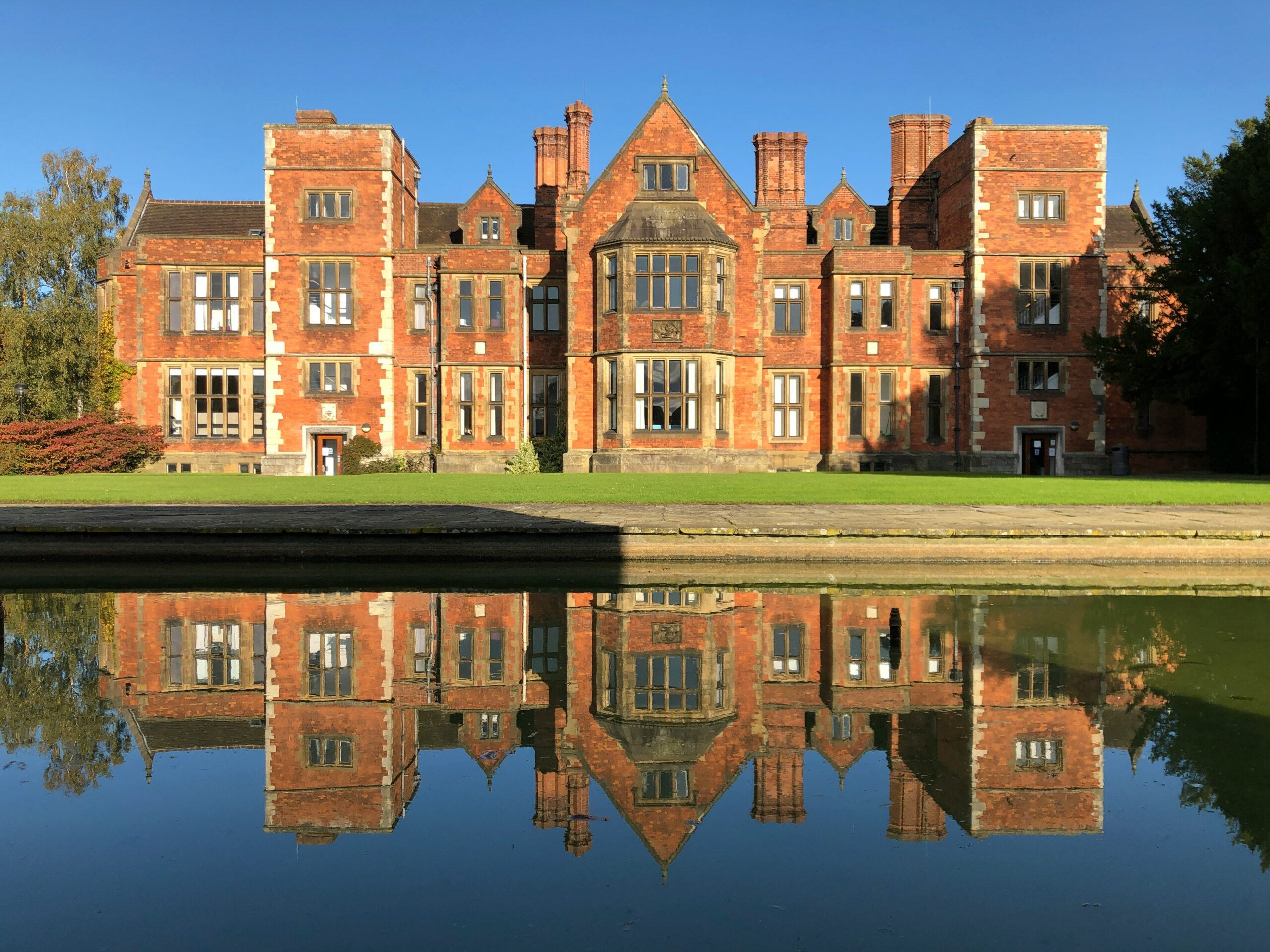 Outside view of a modern University of York building, with its glass façade reflecting beautifully in the calm lake in front. The surrounding greenery and clear sky add to the serene atmosphere, showcasing the university’s integration with nature on its spacious campus.