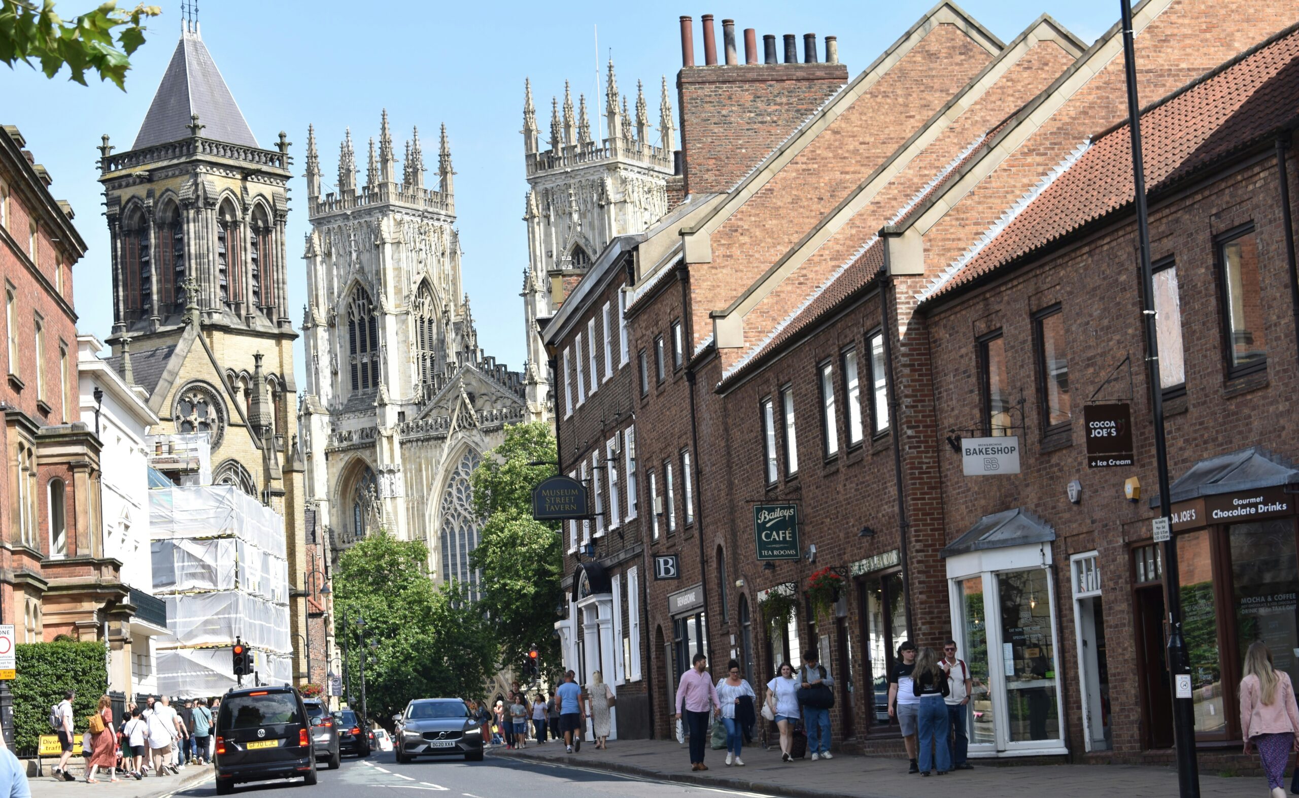 York High Street bustling with pedestrians and historic buildings, leading towards the towering York Minster Cathedral in the background. The Gothic spires of the cathedral rise above the skyline, creating a striking contrast against the quaint medieval architecture of the street.