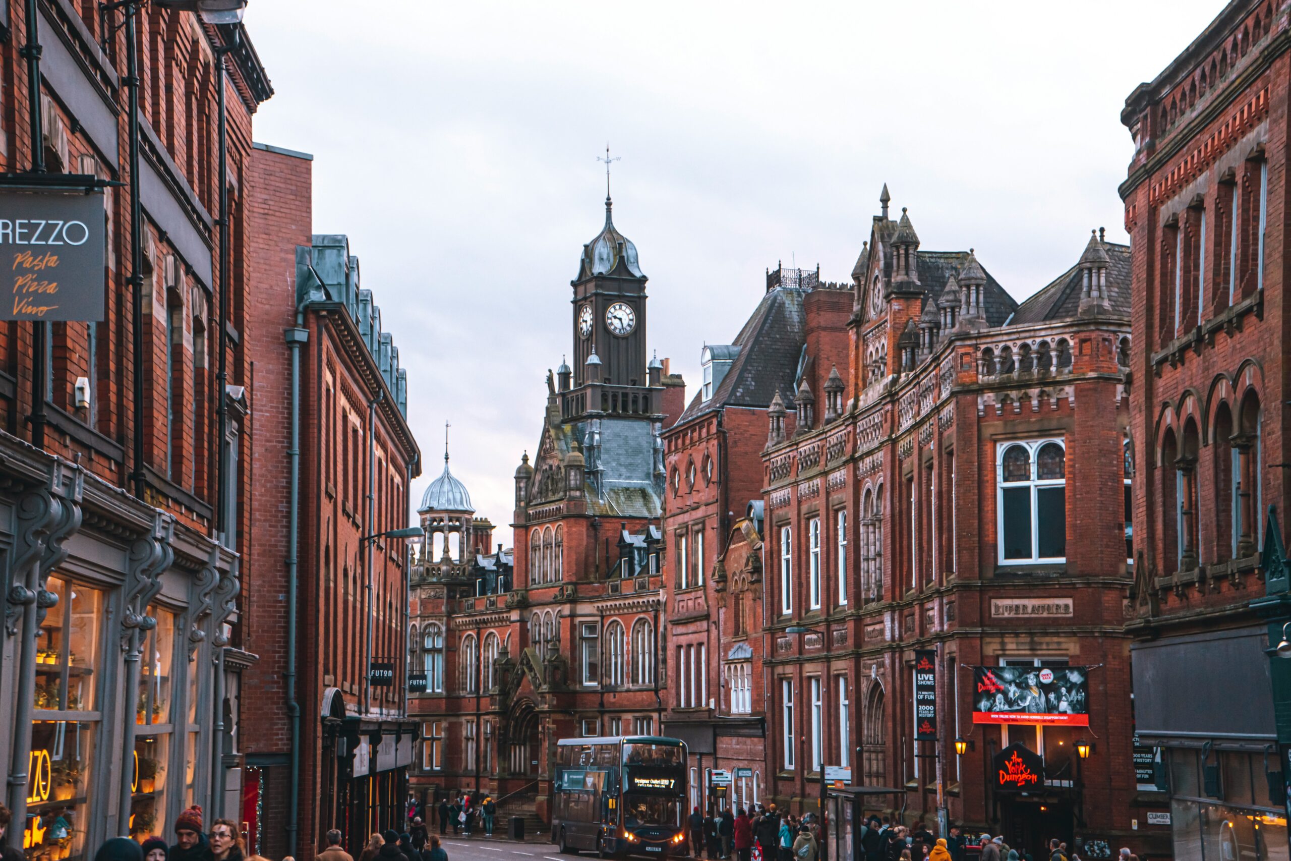 York city centre with its charming cobbled streets, medieval buildings, and vibrant shops. The iconic York Minster towers over the scene in the background, while people stroll through the historic Shambles and other bustling areas filled with cafes and boutiques.