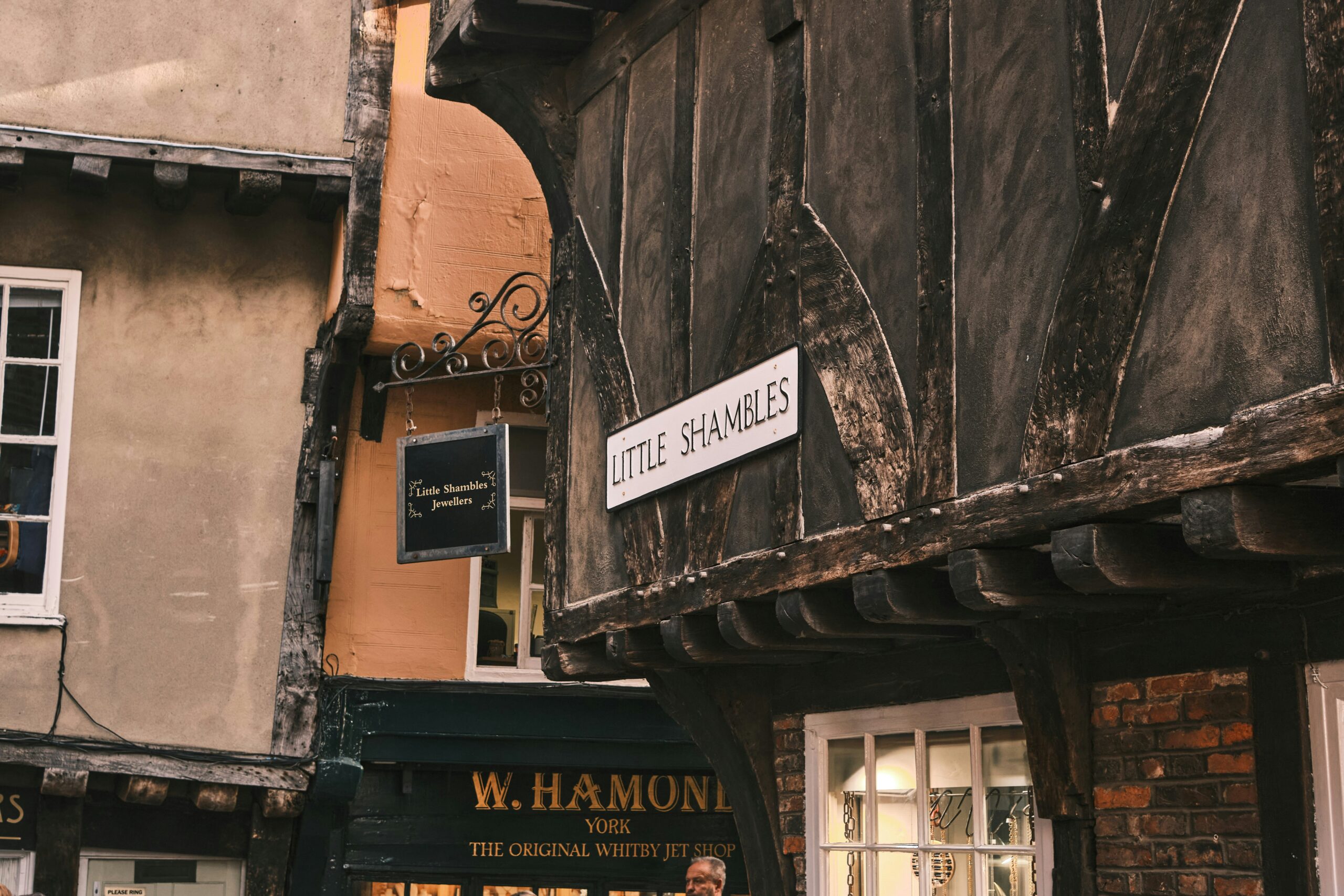 Sign reading 'Shambles' mounted on a historic, timber-framed building in the medieval Shambles street of York, UK. The narrow cobbled street, lined with overhanging buildings, reflects its rich history as a former butcher’s market.